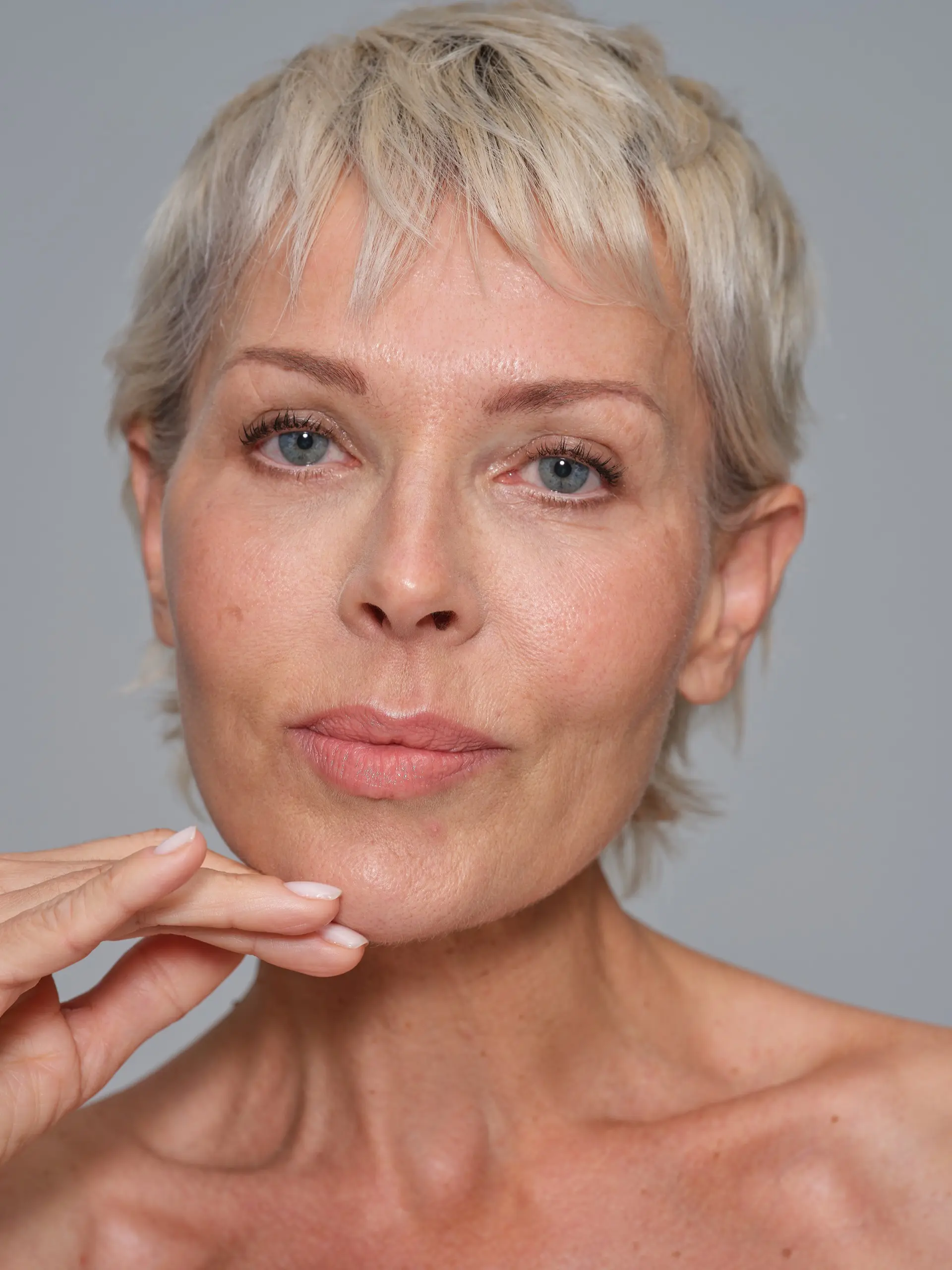 Portrait shot of an mature female model with short blond hair and touching her face in front of a light grey background.