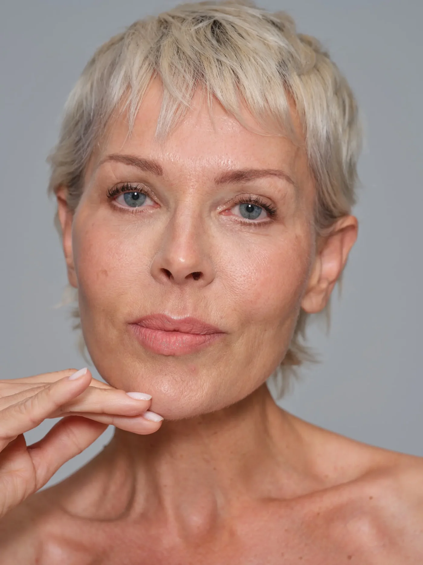 Portrait shot of an mature female model with short blond hair and touching her face in front of a light grey background.